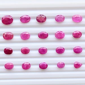 May include: A collection of twenty oval-cut rubies arranged in a four-by-five grid. Each gemstone displays a vibrant pinkish-red hue with faceted surfaces that reflect light, showcasing a variety of sizes and slight color variations.