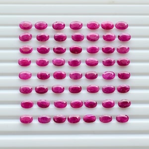 May include: A collection of fifty-six small, oval-shaped, faceted magenta gemstones arranged in an eight-by-seven grid pattern. Each individual stone features a polished surface with multiple facets that reflect light, creating a uniform appearance.
