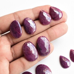 Puede incluir: Una colección de ocho piedras preciosas de rubí facetadas en varias formas, incluyendo ovalada y pera, mostradas en una mano y una superficie blanca. Las piedras preciosas son de un color rojo púrpura profundo y rico.