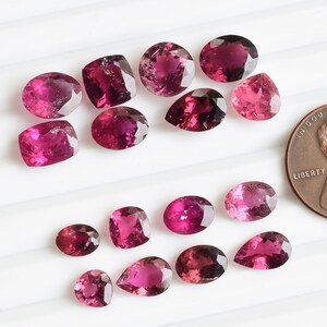 May include: A collection of 15 pink tourmaline gemstones of various shapes and sizes, including oval, pear, and cushion cuts. The gemstones are arranged in a heart shape on a white surface. A US penny is included for scale.