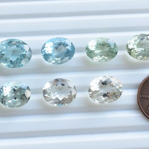 May include: Six oval-shaped gemstones in various shades of green and blue, ranging from light to dark, are arranged in two rows on a white surface. A UK penny is included for size comparison.