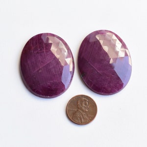 May include: Two oval-shaped, faceted ruby gemstones, each approximately the size of a UK penny.