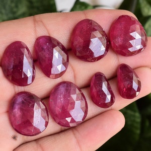 May include: Eight faceted ruby gemstones in various sizes and shapes, displayed on a hand. The gemstones are a deep red color and have a polished finish.