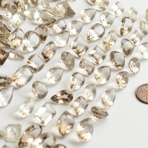 May include: A collection of light brown and clear faceted gemstone beads in various shapes, including pear, square, and rectangular, with a penny for scale.