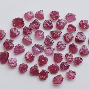 Può includere: Una collezione di circa 40 piccole pietre preziose di rubino rosa grezzo. Le pietre preziose sono sparse su una superficie bianca accanto a un centesimo di euro per la scala.