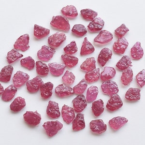 May include: A collection of approximately 40 small, rough, pink ruby gemstones. The gemstones are scattered on a white surface. A penny is included for scale.
