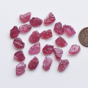 Peut inclure: Une collection de pierres brutes de rubis roses, certaines avec une teinte rougeâtre, éparpillées sur une surface blanche. Un centime d'euro est inclus pour l'échelle.
