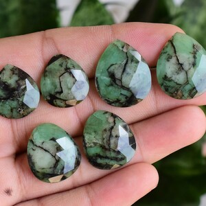 Könnte beinhalten: Sieben grüne und schwarze facettierte tropfenförmige Edelsteine. Die Edelsteine sind in einer Hand angeordnet.