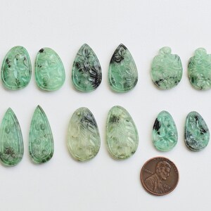 Puede incluir: Una colección de 12 colgantes de piedra preciosa tallada de color verde y negro en forma de lágrima. Los colgantes están dispuestos en dos filas de seis. Se incluye una moneda de un centavo para la escala.