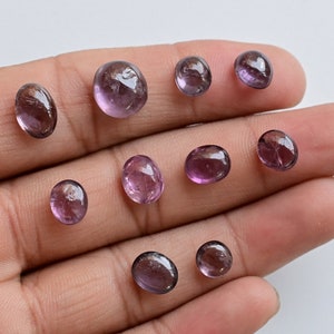 May include: A collection of nine oval-shaped, translucent purple gemstones. The stones are various sizes and have a slight shimmer.