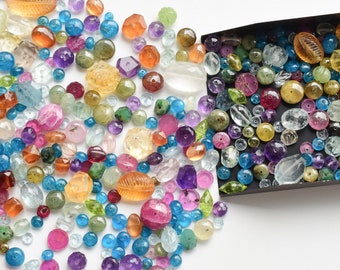 May include: A variety of colorful gemstone beads in various shapes and sizes, including round, oval, and faceted. The beads are scattered on a white surface and spilling out of a black tray.