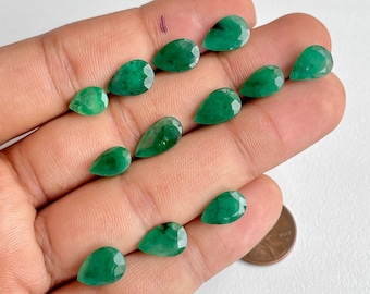 May include: A collection of 12 pear-shaped emerald gemstones, ranging in size, displayed on a hand. The gemstones are a vibrant green color and have a smooth, polished finish.