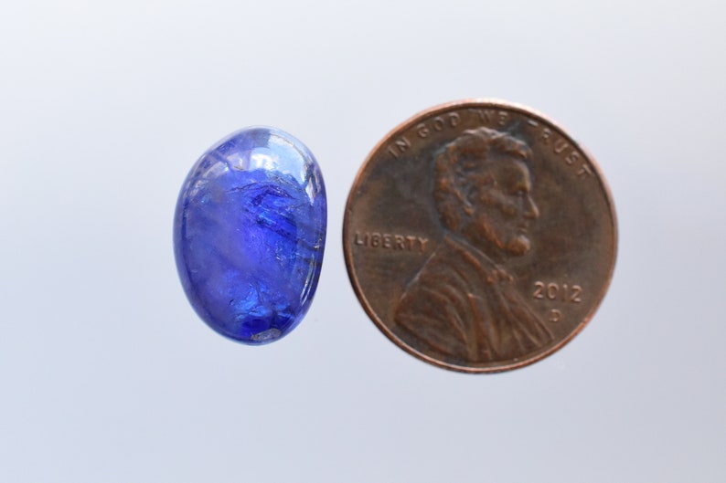 May include: A blue oval gemstone with a faceted surface, next to a US penny for size comparison.