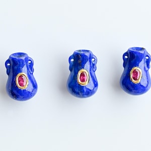 May include: Three blue, vase-shaped pendants with a gold-colored oval and a red gemstone in the center. Each pendant has two small handles near the top. The pendants are displayed on a white background.