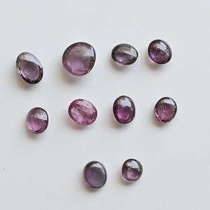 May include: Ten oval-shaped gemstones in shades of purple and pink. The stones are arranged in a random pattern on a white background.