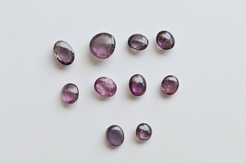 May include: Ten oval-shaped gemstones in shades of purple and pink. The stones are arranged in a random pattern on a white background.