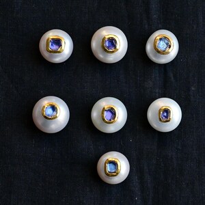 May include: Seven white pearl beads with a gold rim and a square, blue gemstone centre are arranged on a dark background. The pearls have a lustrous sheen, and the gemstones vary slightly in colour.