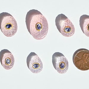 May include: Eight pink, oval-shaped gemstones with a textured surface and a gold-colored bezel. Each stone features a blue, oval-shaped center. A penny is included for size comparison.