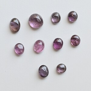 May include: Ten round, faceted, purple gemstones with a slight pink hue. The gemstones are arranged in a loose pattern on a white background.