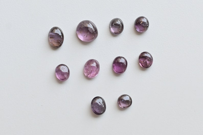 May include: Ten round, faceted, purple gemstones with a slight pink hue. The gemstones are arranged in a loose pattern on a white background.