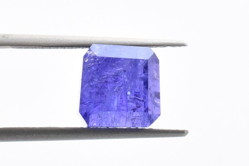 May include: A deep purple gemstone with a rectangular cut, held by tweezers against a white background.