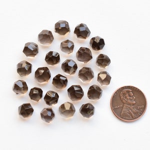K&ouml;nnte beinhalten: Eine Sammlung facettierter Rauchquarz-Edelsteinperlen. Die Perlen sind von transluzenter brauner Farbe und haben eine geometrische Form. Eine M&uuml;nze ist zum Gr&ouml;&szlig;envergleich enthalten. Die Perlen sind auf einer wei&szlig;en Oberfl&auml;che verstreut.