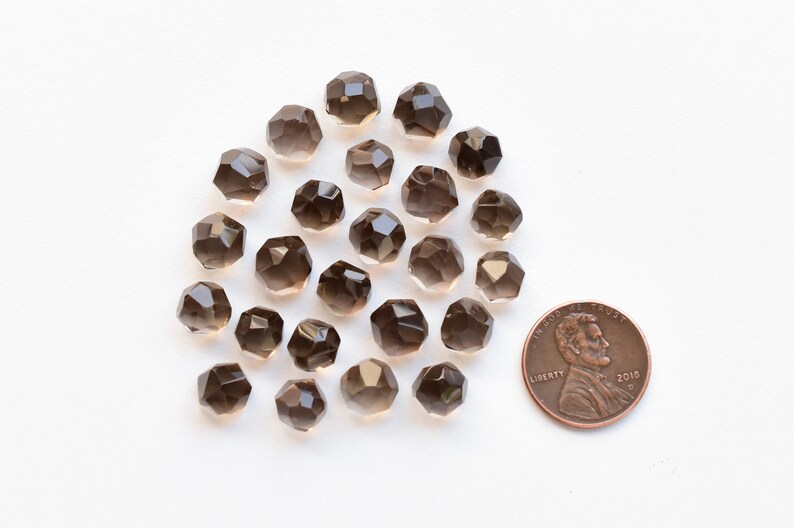 K&ouml;nnte beinhalten: Eine Sammlung facettierter Rauchquarz-Edelsteinperlen. Die Perlen sind von transluzenter brauner Farbe und haben eine geometrische Form. Eine M&uuml;nze ist zum Gr&ouml;&szlig;envergleich enthalten. Die Perlen sind auf einer wei&szlig;en Oberfl&auml;che verstreut.