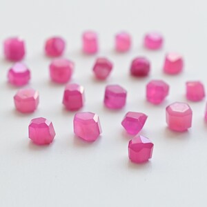 May include: A collection of pink faceted gemstones, scattered on a white surface. The gemstones are various sizes and shapes, with a polished, shiny finish.
