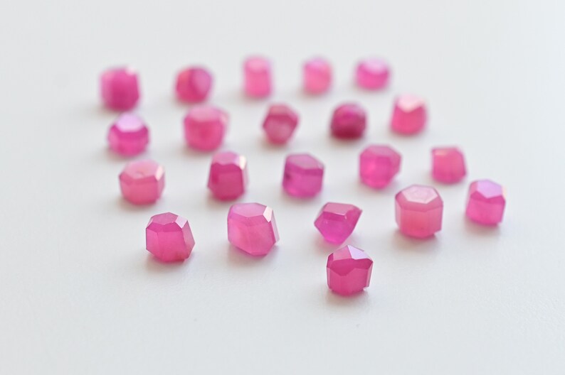 May include: A collection of pink faceted gemstones, scattered on a white surface. The gemstones are various sizes and shapes, with a polished, shiny finish.