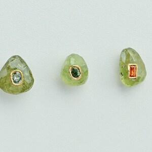 May include: Three olive-green gemstone earrings with gold settings. Each earring features a different colored gemstone: blue-green, dark green, and orange. The earrings are displayed on a white surface.
