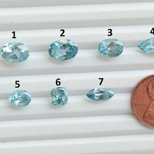 May include: Seven light blue gemstones of various shapes are displayed, including oval, round, and pear-shaped cuts. A US penny is placed next to the gemstones for size comparison. The gemstones are numbered 1 through 7.