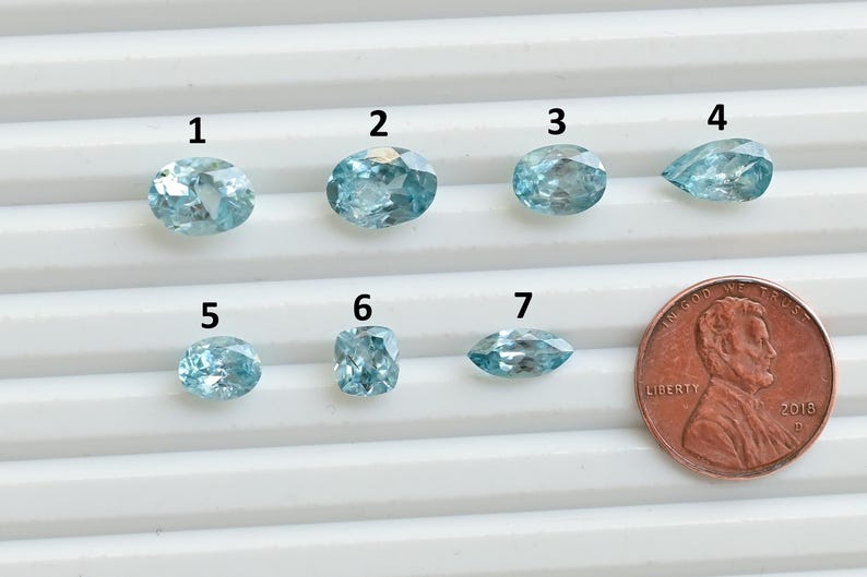 May include: Seven light blue gemstones of various shapes are displayed, including oval, round, and pear-shaped cuts. A US penny is placed next to the gemstones for size comparison. The gemstones are numbered 1 through 7.