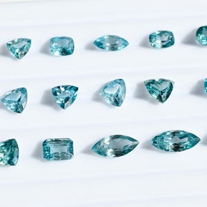 May include: A collection of aquamarine gemstones in various shapes, including round, oval, and square cuts. The gemstones are a light blue color and are arranged on a white surface.