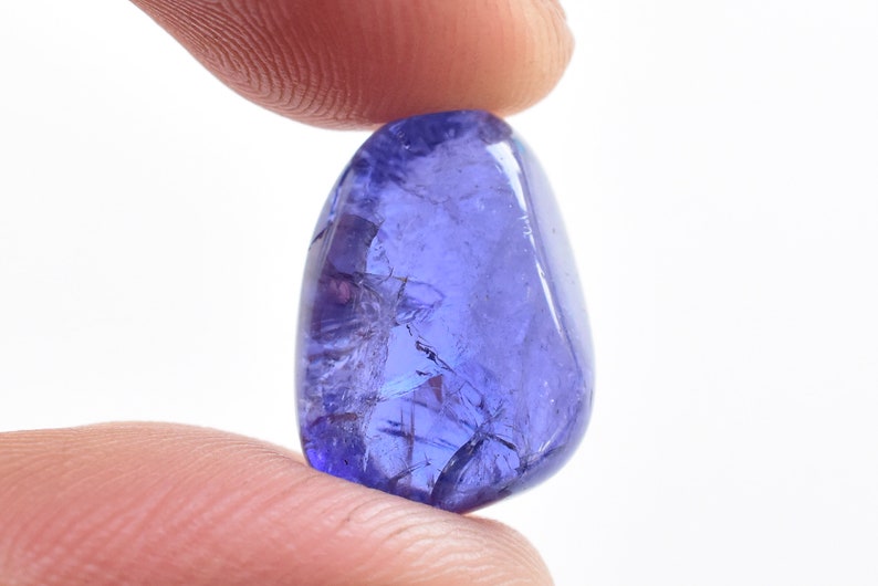 K&ouml;nnte beinhalten: Ein polierter, oval geformter Tansanit-Edelstein, der zwischen zwei Fingern gehalten wird. Der Edelstein hat eine tiefviolett-blaue Farbe mit sichtbaren inneren Br&uuml;chen. Der Hintergrund ist schlicht wei&szlig; und hebt die Farbe und Klarheit des Steins hervor.