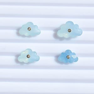 以下が含まれることがあります： 小さな丸い金色の中心部が付いた、4つの水色の雲型のイヤリング。 イヤリングは、白く波状の背景に配置されています。 イヤリングは水色です。