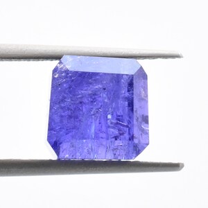 May include: A deep purple gemstone with a rectangular cut, held by tweezers against a white background.