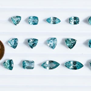 May include: An assortment of teal-colored gemstones in various shapes, including round, square, and marquise cuts, are displayed on a white surface. A US penny is included for size comparison. The gemstones have a sparkling, faceted appearance.