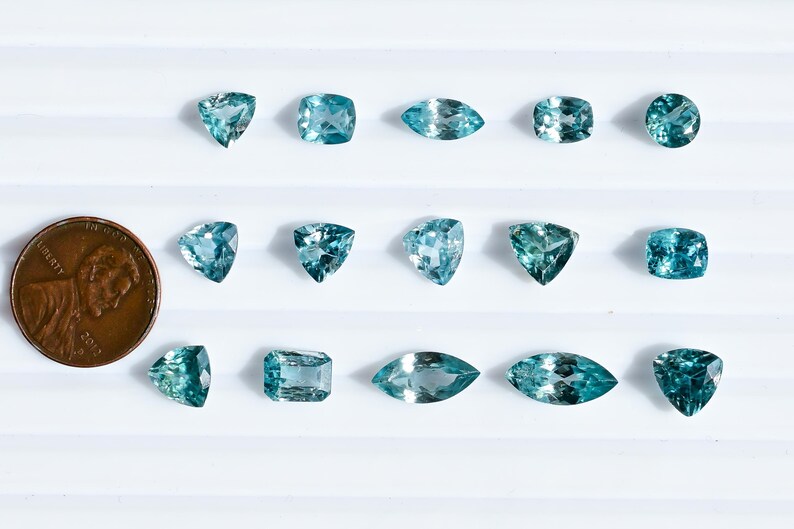 May include: An assortment of teal-colored gemstones in various shapes, including round, square, and marquise cuts, are displayed on a white surface. A US penny is included for size comparison. The gemstones have a sparkling, faceted appearance.
