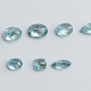 May include: Seven light blue gemstones in various shapes, including oval, round, and pear-cut. The gems are displayed on a white surface, showcasing their clarity and facets. The gemstones are likely for jewelry making.