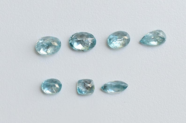 May include: Seven light blue gemstones in various shapes, including oval, round, and pear-cut. The gems are displayed on a white surface, showcasing their clarity and facets. The gemstones are likely for jewelry making.