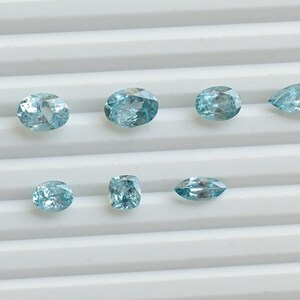 May include: A collection of eight light blue gemstones in various shapes, including oval, pear, and rectangular cuts. The gems are displayed on a white, ridged surface, showcasing their clarity and color.