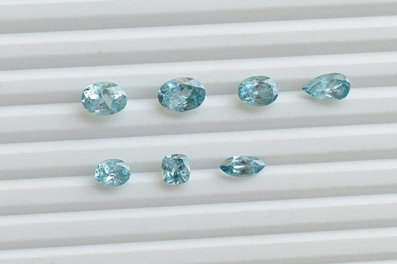May include: A collection of eight light blue gemstones in various shapes, including oval, pear, and rectangular cuts. The gems are displayed on a white, ridged surface, showcasing their clarity and color.