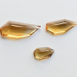 May include: Three faceted, amber-colored gemstones with sharp angles and pointed tips are displayed on a white surface. The gems vary in size, with the largest measuring approximately 1.5 inches in length.