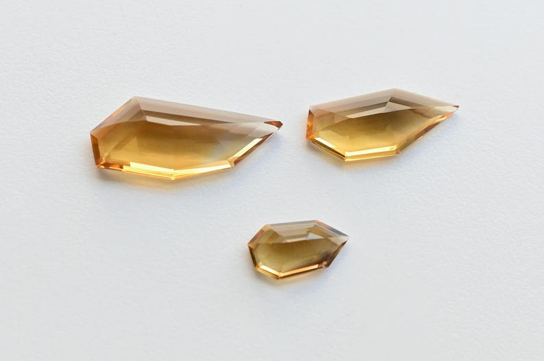 May include: Three faceted, amber-colored gemstones with sharp angles and pointed tips are displayed on a white surface. The gems vary in size, with the largest measuring approximately 1.5 inches in length.