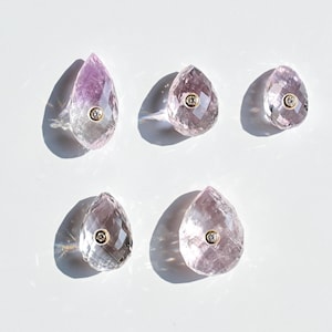 Könnte beinhalten: Fünf facettierte, tropfenförmige Amethyst-Edelsteine mit einem kleinen goldfarbenen Kreis in der Mitte. Die Edelsteine sind auf einer weißen Oberfläche angeordnet, mit Schatten.