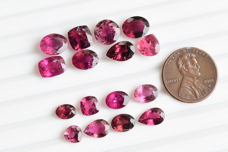May include: A collection of 15 pink tourmaline gemstones of various shapes and sizes, including oval, pear, and cushion cuts. The gemstones are arranged in a heart shape on a white surface. A US penny is included for scale.