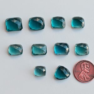 May include: Ten teal blue faceted gemstone cabochons of various sizes, with a UK penny for scale.