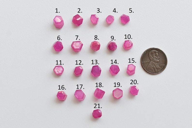 May include: Twenty-one pink faceted gemstones arranged in rows on a white surface. A US penny is included for scale.