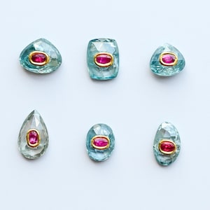 May include: Six faceted gemstones in various shapes, including teardrop, oval, and geometric forms. Each gem features a light blue hue with a vibrant red center stone, set in a gold-colored bezel. The gems are arranged on a white background.
