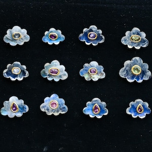 May include: Twelve flower-shaped gemstone pendants with gold-colored settings. The petals are a translucent blue-white, and each pendant features a central gemstone in various colors, including purple, green, and blue. The pendants are arranged on a black surface.
